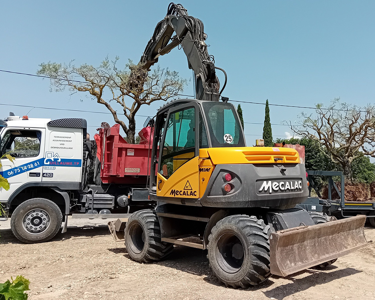 Pelle à roues MECALAC de l'entreprise GUILLAUME TP de Vaison-la-Romaine, en train de charger un olivier dans la remorque du camion benne de GUILLAUME TP.